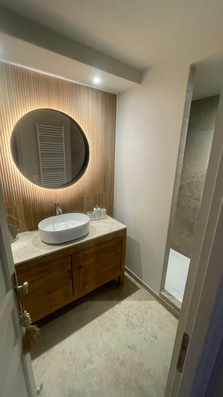 Salle de bain au propre : vasque, miroir rond rétroéclairé et plan en pierre claire